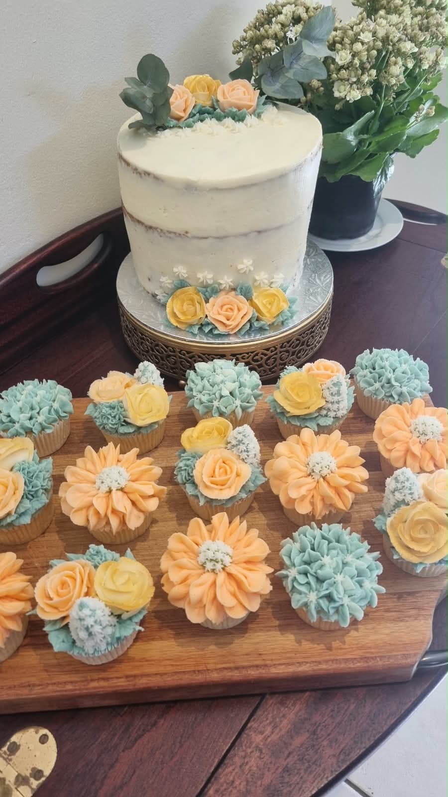 A semi-naked cake with buttercream roses beside peach and teal floral cupcakes